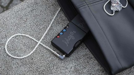 Topview af et lille, sort, bærbart audiokabinet i mat aluminium med indgraveringen "Mojo 2", udstyret med fire farvede, kugleformede knapper og et jackstik forbundet til et sølvfarvet flettet kabel, der fører til gennemsigtige in-ear-hovedtelefoner. Sættet ligger på en grå stenoverflade ved siden af et sort læderetui.