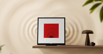 Sort kvadratisk ramme med rød geometrisk illustration centreret på en træhylde, brun lampe til højre, beige væg i baggrunden