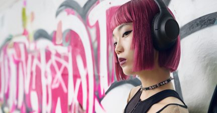 Černé sluchátka na hlavě s růžovými vlasy, zeď s růžovým a černým graffiti v pozadí