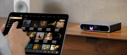 Tīkla audio atskaņotājs ar pelēku anodētu alumīnija korpusu, sānu paneļiem ar ribām, spīdīgu melnu priekšpusi ar divām zeltītām austiņu ligzdām, violetu gaismas apli un krāsu displeju, skatīts no priekšpuses labās puses trīs ceturtdaļu leņķī uz koka mēbeles.
