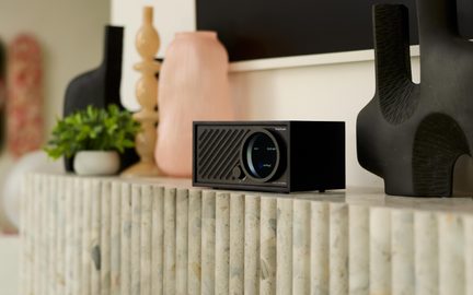 Černý stolní reproduktor, matný kryt z neidentifikovaného materiálu, diagonální mřížka a kulatý displej, pohled ze tří čtvrtin