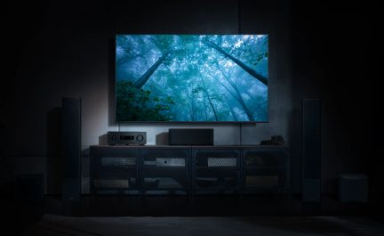 Veggmontert TV som viser en tåkete skog, installert med gulvhøyttalere og svart hjemmekinomøbel