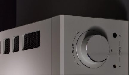 Amplificador de audio plateado con un gran mando selector, dos tomas de auriculares y rejillas de ventilación, visto en ángulo