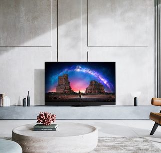 Panasonic TX-65JZ2000E-fladskærm, der viser et billede af et klippelandskab med en regnbue og en person, der står stille, placeret på et lavt betonmøbel i et rum med hvide vægge