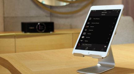 Hvid tablet på metalstativ, der viser en musikmenu med valgmulighederne "This iPhone", "Internet Radio", "Deezer", "Napster", "Qobuz", "Spotify", "TIDAL", "Jazz (We've got)"