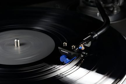 Detailní záběr mírně shora na plastovou černou gramofonovou kazetu s nápisem „NAGAOKA“, vybavenou modrou jehlou, položenou na lesklé černé vinylové desce.