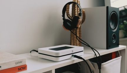 biały odtwarzacz sieciowy Bluesound Node na półce, słuchawki zawieszone na kaktusie i duża szara kolumna hi‑fi obok