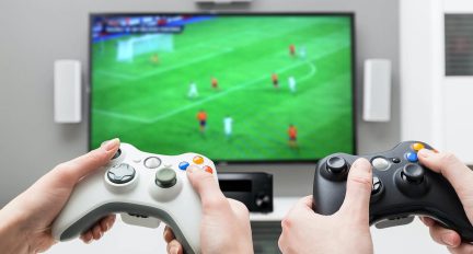 Dvě bílé herní ovladače držené v rukou před televizní obrazovkou zobrazující fotbalový zápas