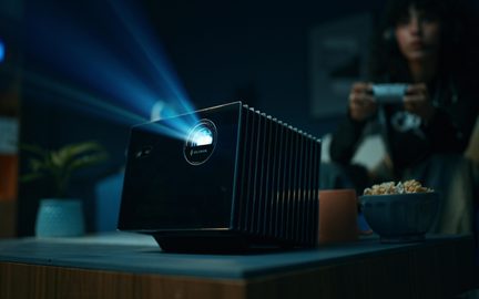 Proiector dreptunghiular negru cu carcasă lucioasă, lentilă frontală iluminată care proiectează o lumină albăstruie și o serie de fante de răcire vizibile pe partea dreaptă, vedere din unghi de trei sferturi față