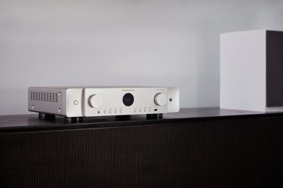 Marantz Cinema 70s gümüş renkli ev sinema sistemi, ön görünüm, koyu renkli ahşap bir mobilya üzerinde duruyor, sağında beyaz bir küp var