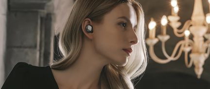 Blondýna z profilu s černými bezdrátovými sluchátky, pohled směrem dolů, rozmazaný lustr v pozadí