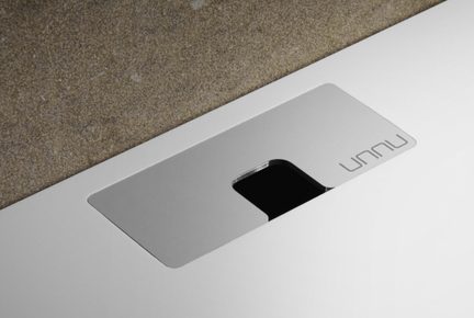 Rektangulær grå metalplade på hvid overflade, U-formet åbning der afslører en sort fordybning, UNNU-logo, skrå visning