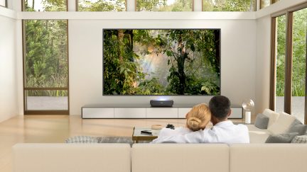 Moderne stue med stor vægmonteret tv, beige sofa, sofabord, panoramavinduer, par sidder med ryggen til