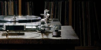 Transparent akryl-pladespiller med klar pladetallerken og chassis, sølvfarvet metal-tonearm og grå cylindriske fødder, set fra siden på et bord af råt træ