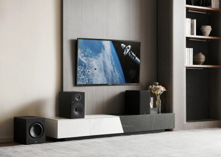 Duvara monte bir ekranın altındaki beyaz ve gri TV ünitesinin çevresinde iki hoparlör ve bir subwoofer içeren siyah ses sistemi