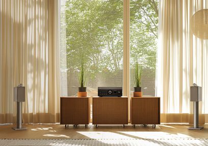Integreret stereo hi-fi forstærker Denon PMA-3000NE på træmøbel, 2 grå højttalere på stativer, 3 potteplanter, beige gardiner