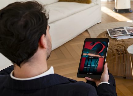 Uomo di spalle in abito che tiene un tablet con un'immagine colorata, seduto su un divano bianco, tavolino in rattan