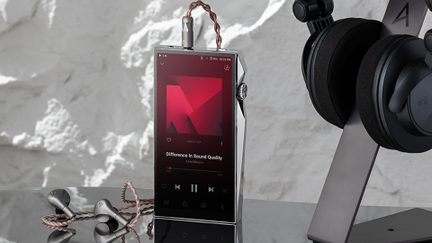 Vista frontale leggermente angolata di un lettore audio digitale rettangolare in metallo argentato lucido con ampio schermo touch nero acceso, rotella zigrinata laterale e jack per cuffie superiore collegato a un cavo intrecciato in rame
