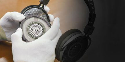 Detailní šikmý záběr černých oboustranných sluchátek s kruhovým kovovým grilováním, pěnovými polstrováním a viditelným šedým vnitřním měničem, drženo v bílých rukavicích