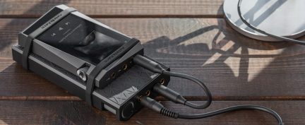 Lettore audio portatile rettangolare nero con schermo, pulsanti visibili, cavi collegati, posizionato su una superficie in legno marrone.