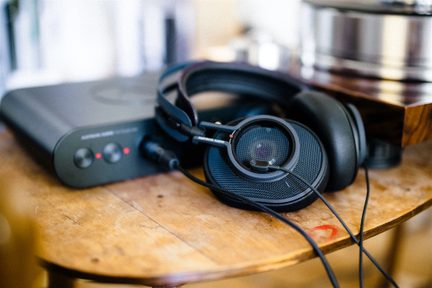 Căști audio cu fir, negre, din plastic, cu grile metalice circulare și pernițe groase din piele ecologică, așezate oblic pe o masă de lemn lângă o carcasă audio neagră, vedere de aproape din unghi oblic
