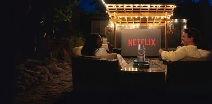 Due persone sedute su un divano in rattan, schermo Netflix, proiettore, pergola in legno con ghirlande luminose