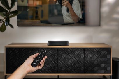 Polk Audio MagniFi Mini AX soundbar i sort, sort fjernbetjening, lyst træ TV-møbel, hånd der holder fjernbetjeningen