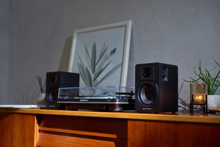 Černá vinylová deska s průhledným víkem, dvě černé reproduktory, rámeček s rostlinnou ilustrací, zapálená svíčka
