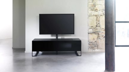 Černá plochá televize na černém nízkém nábytku, bílá zeď vlevo, kamenná zeď vpravo, šedá podlaha, tmavý sloup