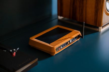 Carcasă multimedia Wi-Fi Orange dreptunghiulară, cu antenă și porturi RCA pe spate, așezată pe un birou albastru