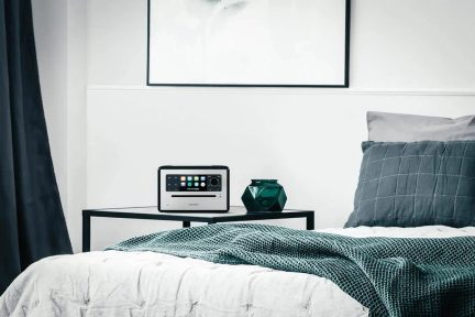 Schlafzimmer mit Bett, blauer gemusterter Tagesdecke, grauen Kissen, schwarzem Nachttisch, Radiowecker, grüner Vase, schwarzem Bilderrahmen an der Wand
