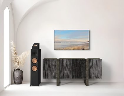 Černý sloupový reproduktor s měděnými měniči a modulem Atmos před TV stolkem v moderním obývacím pokoji