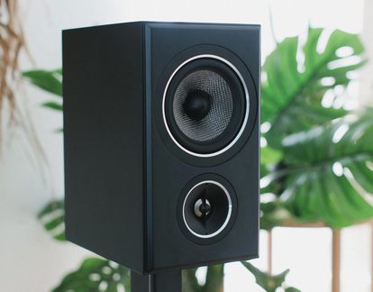 Caixa de som biblioteca preta fosca, vista em três quartos, com woofer de cone tecido e tweeter, arremates metálicos prateados