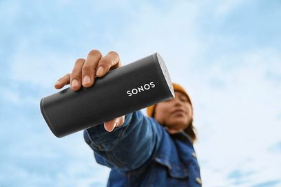 Sonos Roam Shadow Black bærbar Bluetooth-højttaler holdt i en hånd, set forfra, mod en lys blå himmelbaggrund