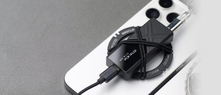 Kleine rechthoekige zwarte matte metalen behuizing met USB-C-poort en jack-uitgang, vastgehouden door een elastisch koord aan een magnetische ring, alles diagonaal geplaatst op de achterkant van een witte smartphone, van bovenaf gezien