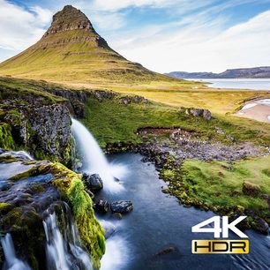Bjergmassiv med vandfald, der løber ned i en snoet flod gennem en grøn dal under en blå, let overskyet himmel