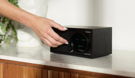 Rádio z černého dřeva s kulatým displejem zobrazujícím Spotify a otočným knoflíkem, pohled ze tří čtvrtin, ruka jej nastavuje