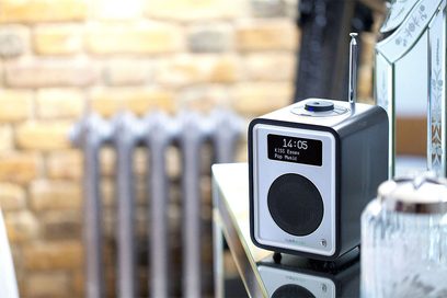Digital radio Ruark Audio R1 Mk3 i metalgrå, LCD-skærm der viser "14:05 KISS Essen Pop Music", placeret på en støbejernsvarmekilde