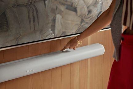 Soundbar Sonos bianca sotto televisore a parete, comandi touch sfiorati da una mano femminile