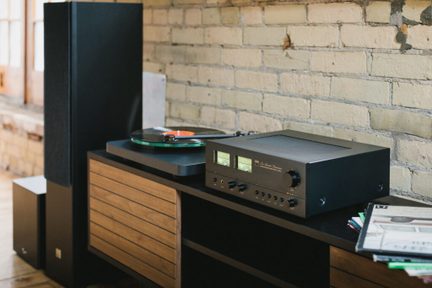 Hi-fi ķēde ar melnu stereo pastiprinātāju un vinila atskaņotāju uz koka TV skapīša ķieģeļu sienas priekšā