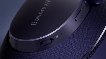 Nahblaufnahme einer Bowers & Wilkins Kopfhörermuschel in Mitternachtsblau mit satiniertem Aluminium und Mesh-Gewebe, schräger Blickwinkel, schwarzer Schiebeschalter mit Netzsymbol, sichtbare grüne LED und Bluetooth-Piktogramm, schmaler bündiger Knopf an der Krone, Belüftungsschlitz auf der Rückseite