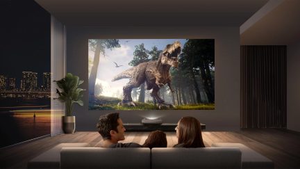 Moderne stue med kæmpe skærm, der viser en dinosaur, 3 personer set bagfra på en grå sofa, grøn plante i potte