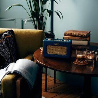 Blå rektangulær bærbar radio i plast med retrofinish, indbygget gylden metalgitter, sølvfarvede knapper på toppen, kromhåndtag på siden og tynd antenne, set fra trekvart forfra