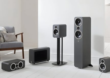 Set of five anthracite gray speakers in a matte wood cabinet, front view, including a tall floorstanding speaker, a bookshelf speaker on a black metal stand, a three-way horizontal center speaker, a rectangular subwoofer with a black grille, and a small bookshelf speaker, all equipped with metal-rimmed drivers