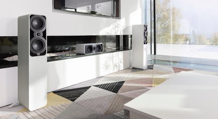 Two white painted wooden floorstanding speakers and their matching center speaker, with black front panels featuring two circular drivers and a central tweeter, shown in perspective in a bright modern living room