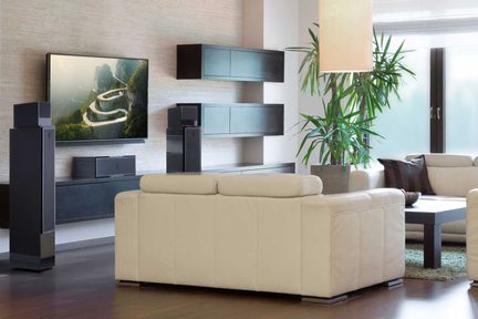 En beige sofa, et vægmonteret fjernsyn, to sorte højttalere, væghylder, en grøn plante, et sofabord