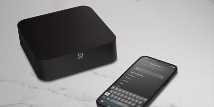 Black Bluesound wireless audio hub with rounded corners, three-quarter view, with setup smartphone