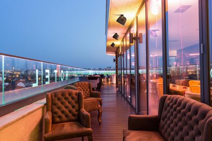 Terrazza rooftop d'hotel con poltrone imbottite marroni, vetrate e vista urbana al crepuscolo
