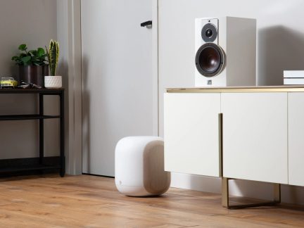 Bílý bezdrátový subwoofer Bluesound PULSE SUB+ se zaoblenými hranami, umístěný na podlaze v obývacím pokoji