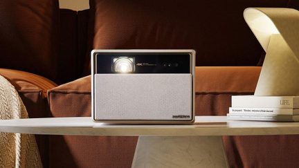 Vit 4K-projektor med Harman Kardon-högtalare med tygfront på soffbord, mysigt vardagsrum med lampa och böcker